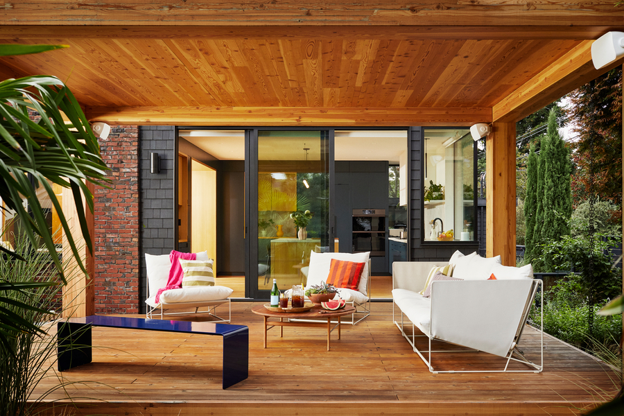 A stylish covered outdoor patio features modern white lounge seating with colorful pillows, a wooden coffee table with drinks and snacks, and a sleek navy bench. White Sonos outdoor speakers are installed on the roof.