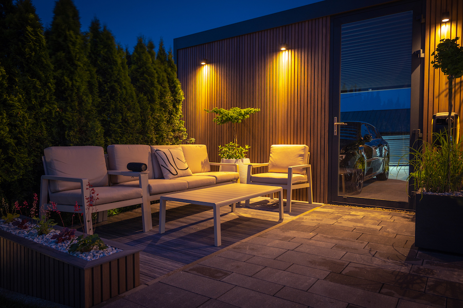 An outdoor patio at night with cushioned chairs positioned around a table. Downlights shine on this display. Next to the patio is a carport with a car visible through the glass door.